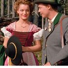 August-Kalenderblatt. Ein Mann und eine Frau in traditioneller Kleidung im Wald. Sie lächeln einander an.