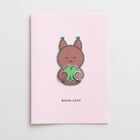 Text: "GOOD LUCK". Illustration: Ein braunes, lächelndes Tier mit großem Kleeblatt auf rosa Hintergrund.