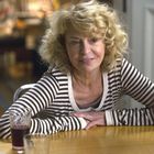 Ich kann keine Texte im Bild sehen. Eine Frau mit lockigem, blondem Haar sitzt an einem Tisch mit Getränken.