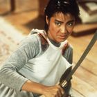 Eine Person mit kurzem Haar hält entschlossen ein Schwert. Hintergrund zeigt Holzboden und Teppich.