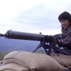 Eine Frau in einer Jacke bedient ein schweres Maschinengewehr, gestützt auf Sandsäcke vor einem Berg- und Wiesenhintergrund.