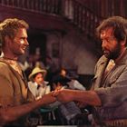 Zwei Männer stehen sich gegenüber und halten Hände. Im Hintergrund eine Treppe und unscharfe, sitzende Personen.