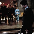 Ein Mann im Anzug steht vorn, schaut nach unten. Mehrere Personen, darunter eine Frau im Kimono, gehen im Hintergrund.