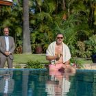 Kein Text sichtbar. Ein Mann in rosa Shorts sitzt am Pool, ein anderer steht dahinter, beide umgeben von Pflanzen.