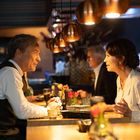 Zwei Personen unterhalten sich an einem Restauranttisch bei warmem Licht; Blumen und Geschirr sind auf dem Tisch.