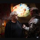 Zwei Männer stehen in einem düsteren Raum. Einer trägt schwarze Kleidung, der andere eine Rüstung; eine bunte Glasdecke im Hintergrund.