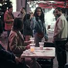 Eine Frau grüßt lächelnd, während Menschen an einem Straßenimbiss stehen und andere an einem Tisch sitzen.