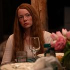 Eine Frau mit langen roten Haaren sitzt an einem Tisch, umgeben von Tellern und einem Blumenstrauß mit rosa Tulpen.