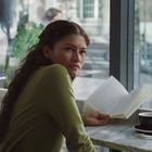 Eine Frau mit langen Haaren sitzt in einem Café, hält ein Buch und schaut aus dem Fenster; eine Tasse Kaffee steht auf dem Tisch.