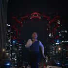 Eine Person rennt bei Nacht durch eine belebte Stadt, Gebäude und Lichter umgeben sie. Ein großes, leuchtendes, rot-schwarzes Tor.