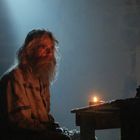 Ein älterer Mann mit langem Haar und Bart sitzt in einem schwach beleuchteten Raum neben einer Kerze.