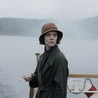 Eine Frau mit braunem Hut und grünem Mantel steht auf einem Schiff, hält eine Zigarette und schaut zur Seite. Im Hintergrund Wasser.