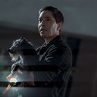 Ein Mann hält einen Hund im Arm und eine Taschenlampe in der Hand, wirkt besorgt. Es ist dunkel und leicht regnerisch.