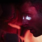 Kein Text sichtbar. Ein Mann mit ernstem Ausdruck, rotes Licht beleuchtet sein Gesicht seitlich.