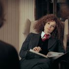 Eine Frau mit lockigem Haar in einem Anzug sitzt nachdenklich in einem Sessel und hält ein Notizbuch.