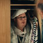 Ein Mann in einem Schrank trägt einen weißen Hut und Pelzmantel. Ein anderer Mann schaut hinein, während bunte Kleidung hängt.