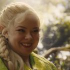 Ich kann die Texte oder Personen nicht identifizieren. Junge Frau mit blondem Zopf in grünem Gewand, lächelnd im Wald.