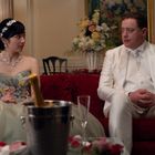 Ein Paar in eleganter Kleidung sitzt auf einem roten Sofa. Vor ihnen steht ein Eiskübel mit einer Champagnerflasche.