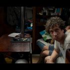 Zwei Männer in einem gemütlichen Raum, einer blickt nachdenklich, der andere sitzt ohne Hemd.