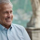 Es gibt keinen Text. Ein Mann im hellblauen Hemd lächelt vor einer verschwommenen, grünen Landschaft mit Wasser im Hintergrund.