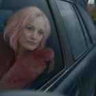 Ich kann den Text nicht lesen. Eine Frau mit rosa Haaren schaut aus einem Autofenster. Straße und Bäume im Hintergrund.