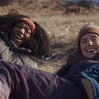 Zwei Menschen lachen, während sie in warmer Kleidung auf einer Wiese liegen. Die Stimmung wirkt fröhlich und entspannt.