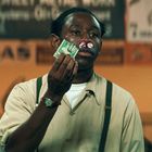 Ein Mann hält Geldscheine in der Hand, hat eine blutige Nase mit Watte, trägt ein Hemd mit Hosenträgern. Im Hintergrund sind unscharfe Schilder.