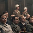 Ich kann den Text nicht lesen. Es sind Männer in Uniformen und Anzügen, die in einem Gerichtssaal sitzen.
