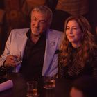 Ein Mann in hellem Sakko und eine Frau sitzen lächelnd in einem Club; er hält ein Getränk.