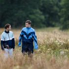 Zwei Menschen in blauen Trainingsanzügen spazieren durch eine Wiese im Regen. Bäume im Hintergrund.