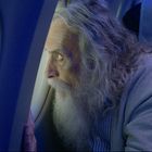 Text: "Airlines Vero". Ein älterer Mann mit langem, grauem Haar und Bart schaut aus einem Flugzeugfenster.