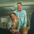 Zwei Personen mit Gläsern, eine sitzt lächelnd auf dem Sofa. Weitere Personen im Vordergrund sind unscharf zu erkennen.