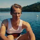 Ein junger Mann mit Brille im Ruderboot auf einem See, trägt ein Trikot mit olympischen Ringen. Hintergrund: bewaldete Küste.