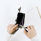 Zwei Hände formen mit einer Zange ein schwarzes Papierobjekt, das Draht enthält, auf einem weißen Hintergrund.