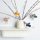 Es gibt keine Texte. Eine Vase mit Zweigen und bunten Filz-Schmetterlingen auf einem weißen Regal.