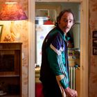 Ein Mann in Trainingsjacke, mit Krücke, steht in einem gemütlichen, leicht unordentlichen Raum mit Stehlampe und gerahmten Fotos.