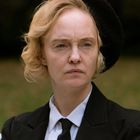 Eine Person mit ernstem Gesichtsausdruck trägt einen schwarzen Anzug und Hut vor verschwommenem, grünen Hintergrund.