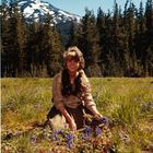 Eine lächelnde Frau sitzt auf einer Wiese mit violetten Blumen, im Hintergrund ein schneebedeckter Berg und viele Tannenbäume.