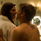 Ein Mann und eine Frau lächeln sich in einem Badezimmer an. Sie trägt einen Bademantel. Ein beleuchteter Spiegel ist im Hintergrund.