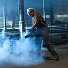 Eine Person steht in einer Fabrikhalle und hält eine Kettensäge, umgeben von Rauch oder Dunst.