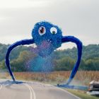 Ein blaues, flauschiges Wesen mit großen Augen sprüht farbigen Staub. Eine Person läuft auf einer Straße davor weg.