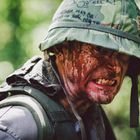 Auf dem grünen Helm steht: "Vietnam may be hazardous to your health." Ein Soldat schaut grimmig, mit Tarnkleidung.