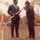 Ein Mann im Anzug hält ein Brett und steht vor einem Polizisten im Freien, umgeben von herbstlichen Bäumen und einem Bagger.