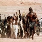 Eine Frau in weißem Kleid geht neben Männern mit traditionellen Schilden und Speeren durch eine Wüstenlandschaft.