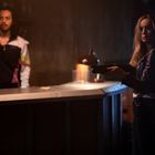 Eine Frau in Lederjacke hält ein Gewehr in einer dunklen Bar. Ein Mann im Sportanzug steht hinter der Theke.