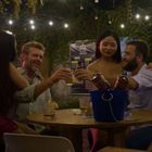 Ein Menu-Poster hängt im Hintergrund. Vier Personen sitzen an einem Tisch und stoßen mit Getränken unter Lichterketten an.