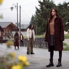 Im Zentrum stehen fünf Personen auf einer Dorfstraße, ernst wirkend, mit Bäumen und Häusern im Hintergrund.