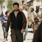 Eine Person mit Bart und dunkler Kleidung geht in einer belebten Straße. Menschen bewegen sich im Hintergrund umher.