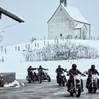 Keine sichtbaren Texte. Fünf Motorradfahrer fahren auf verschneiter Straße, schneebedeckte Hügel im Hintergrund.