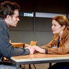 Zwei Personen sitzen sich an einem Tisch gegenüber. Sie halten Händedesign eine ernste Atmosphäre ausdrückt. Eine Frau trägt eine braune Jacke.